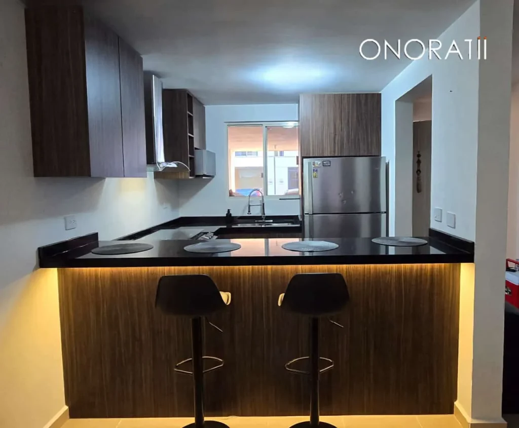 Una barra desayunadora que es de color oscuro, tiene dos sillas enfrente que también son de colo oscuro, la barra tiene una iluminación por debajo, se ve que es luz amarilla, los gabinetes que hay en la pared combinan con la base de la barra, está en una habitación blanca